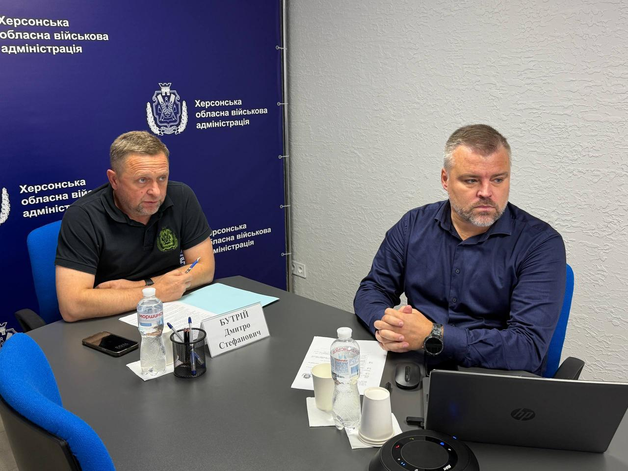 Перший заступник начальника Херсонської ОВА Дмитро Бутрій та директор Департаменту розвитку економіки ХОВА Артем Мішин сидять за робочим столом під час онлайн-зустрічі в межах платформи «Діалог влади та бізнесу», присвяченої підтримці підприємств. Ліворуч на передньому плані сидить Дмитро Бутрій у чорній футболці-поло з тактичним шевроном, який робить нотатки ручкою на бланках поруч із настільною табличкою з власним ім'ям та пляшкою води. Праворуч від нього зафіксований Артем Мішин у темно-синій сорочці, який уважно слухає доповідачів, тримаючи руки разом біля відкритого ноутбука HP та круглого чорного спікерфона. Нарада проходить на тлі великого темно-синього офіційного банера з чисельними повтореннями герба ХОВА та білими написами «Херсонська обласна військова адміністрація», що ілюструє обговорення змін у програмі компенсацій за пошкоджене майно та оновлень у Міжнародному реєстрі збитків (RD4U).