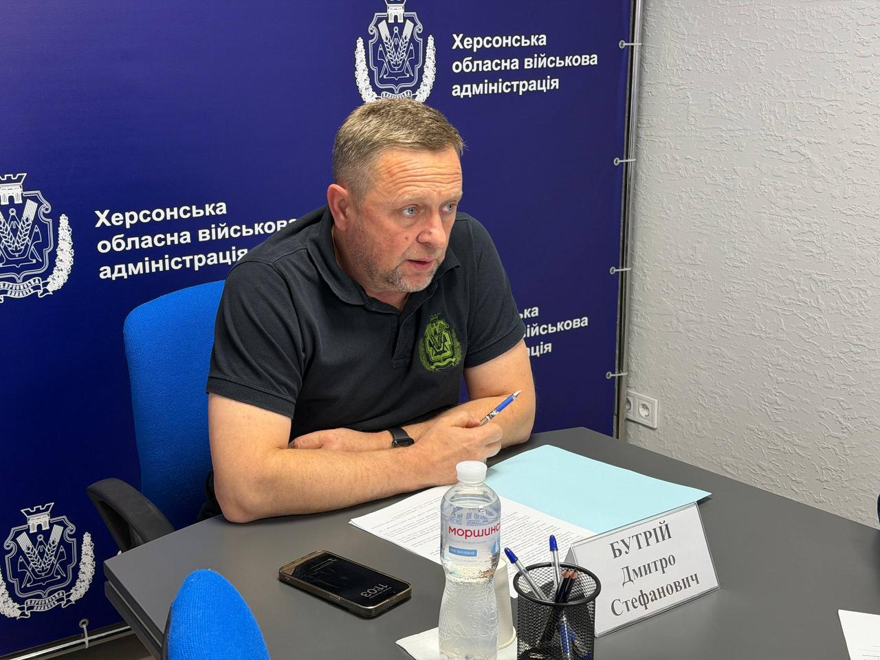Перший заступник начальника Херсонської ОВА Дмитро Бутрій сидить за робочим столом у кабінеті під час онлайн-зустрічі в межах платформи «Діалог влади та бізнесу», присвяченої підтримці підприємств. Посадовець у чорній футболці-поло з тактичним зеленим шевроном на грудях зафіксований крупним планом, коли він веде діалог, тримаючи в руці синю ручку перед розкладеними робочими паперами. На сірому столі перед ним лежать мобільний телефон, пляшка води «Моршинська», підставка з канцелярією та встановлено білу настільну табличку з написом «БУТРІЙ Дмитро Стефанович». Зустріч проходить на тлі великого темно-синього офіційного банера з чисельними повтореннями герба ХОВА та білими написами «Херсонська обласна військова адміністрація», що ілюструє обговорення оновлень у Міжнародному реєстрі збитків (RD4U) та програм компенсації для постраждалого від війни бізнесу.