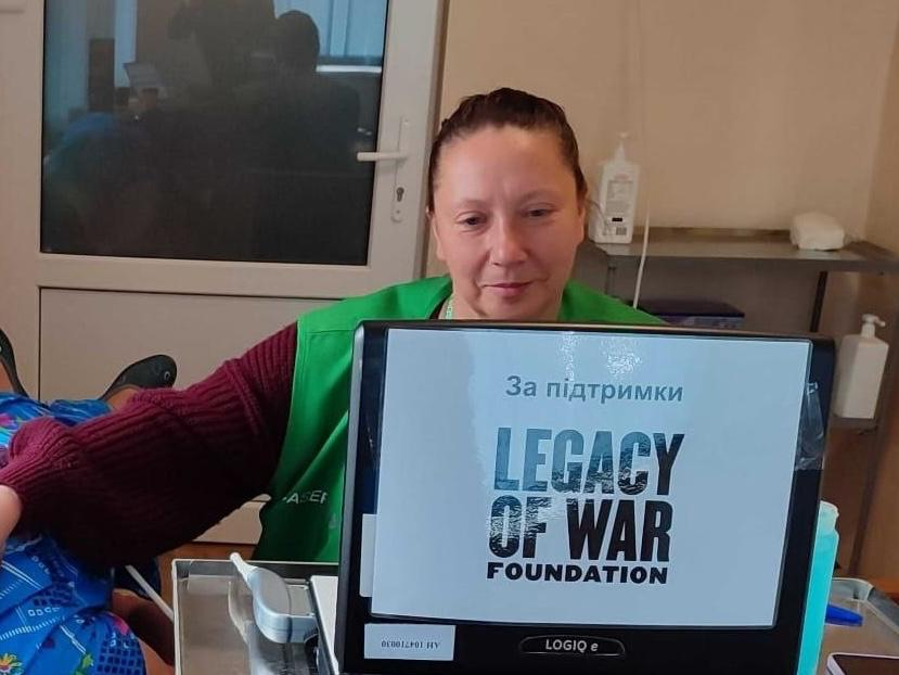Лікарка мобільної медичної команди «Legacy of War Foundation» у зеленому фірмовому жилеті сидить перед монітором портативного апарату УЗД LOGIQ e під час проведення ультразвукового обстеження пацієнта. Фахівчиня у бордовому светрі зафіксована за робочим столом у медичному кабінеті, а ліворуч на кушетці видно ноги пацієнта під синім кольоровим простирадлом. На чорному екрані діагностичного обладнання відображається фірмова заставка з написом «За підтримки LEGACY OF WAR FOUNDATION», що ілюструє проведення діагностичних обстежень на Херсонщині, де за п'ять днів 145 мешканців громад безкоштовно отримали фахову допомогу невролога та спеціаліста з УЗД.