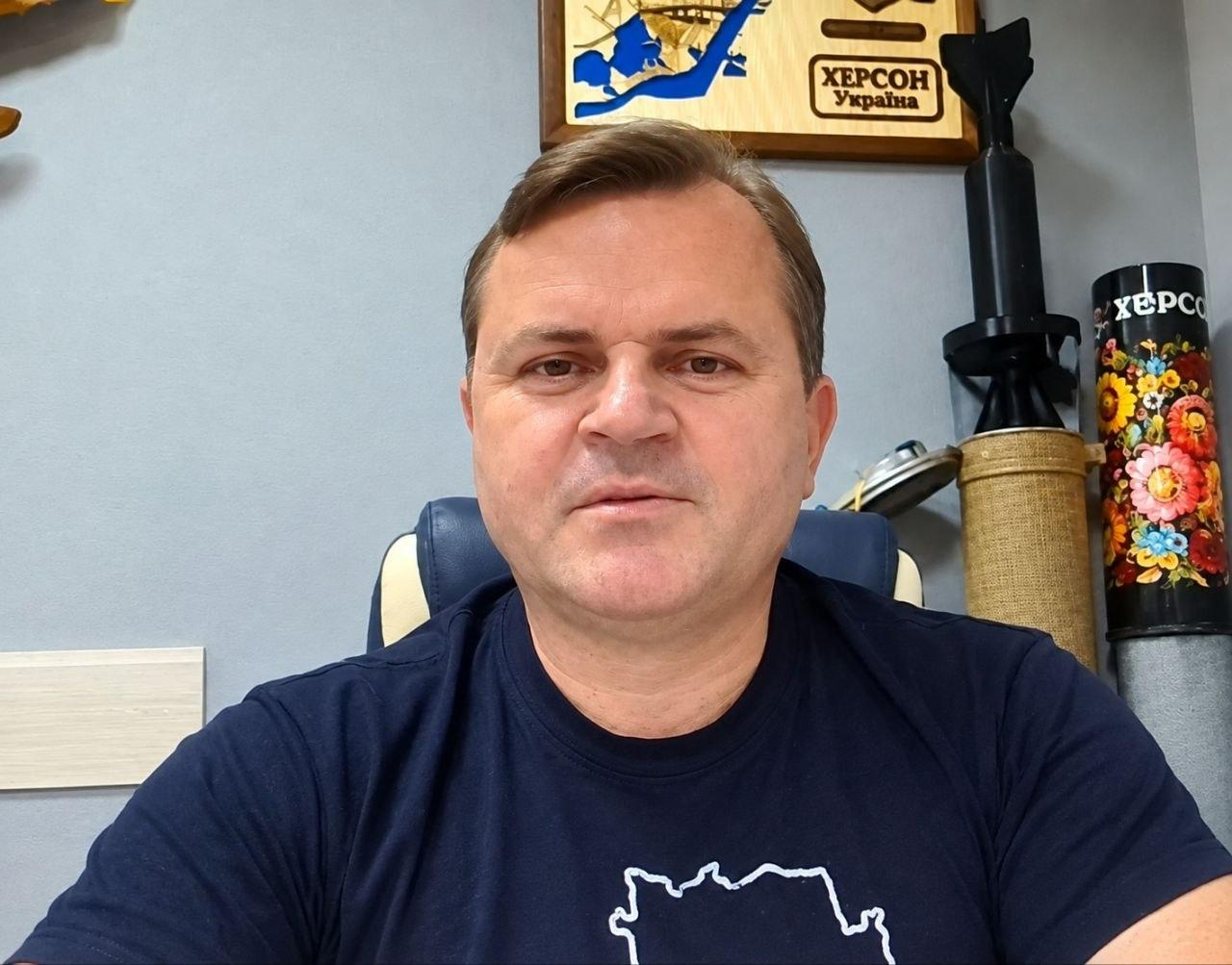 Заступник начальника Херсонської ОДА Володимир Клюцевський зафіксований крупним планом в офісному кріслі під час головування на черговому засіданні гуманітарного штабу ХОВА у форматі онлайн-конференції. Посадовець у темно-синій футболці з контурним білим малюнком карти України дивиться в камеру на тлі світло-сірої стіни кабінету. На задньому плані над його плечима розміщено декоративне дерев'яне панно у вигляді мапи Херсонської області з написом «ХЕРСОН Україна», а праворуч на полиці встановлено військові сувеніри, тубус та гільзу, прикрашену яскравим петриківським розписом і написом «Херсон». Кадр ілюструє проведення робочої наради, під час якої ухвалили рішення щодо забезпечення медичних закладів резервними джерелами живлення і водою, а також безперебійної роботи системи оповіщення у Херсоні.