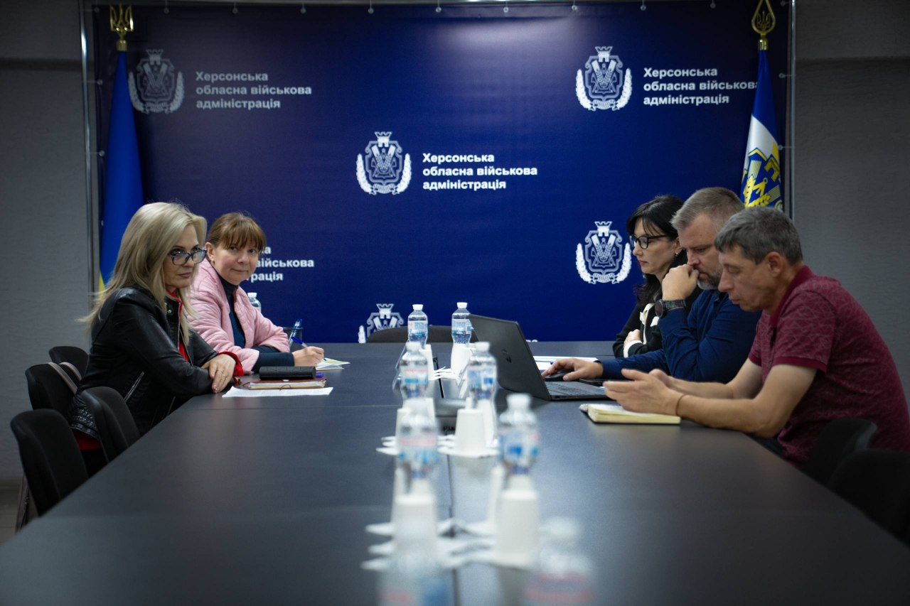 Учасники наради сидять за довгим робочим столом у залі засідань на тлі великого темно-синього фірмового банера з гербами ХОВА та білими написами «Херсонська обласна військова адміністрація» під час онлайн-підключення до засідання Національної Ради безбар’єрності. Ліворуч за столом спочатку сидить директорка Департаменту здоров’я Херсонської ОДА Тетяна Карчевич у рожевій куртці, а поруч із нею — колега у чорній шкіряній куртці та окулярах. Праворуч розташовані троє учасників зустрічі, серед яких директор Департаменту розвитку економіки Херсонської ОДА Артем Мішин у синій кофті працює за відкритим ноутбуком, а поруч у напівпрофіль показаний начальник управління фізичної культури, молоді та спорту Херсонської ОДА Сергій Яворський у бордовій футболці-поло, який робить нотатки у блокноті. Загалом кадр відображає участь представників адміністрації у всеукраїнському обговоренні реалізації державної політики, за підсумками якого Херсонщина посіла четверте місце в загальноукраїнському рейтингу виконання Плану заходів із безбар’єрності.