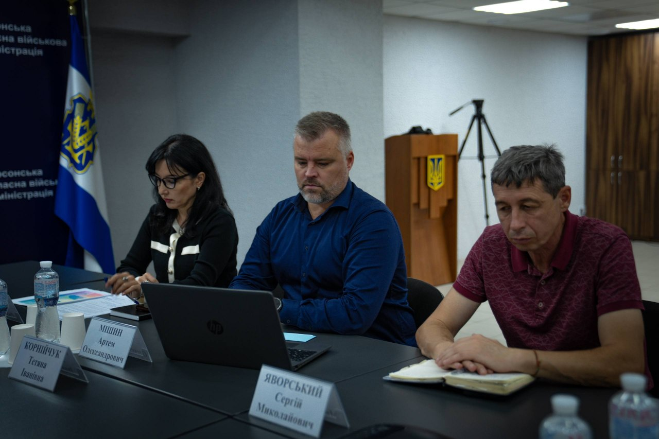 Директор Департаменту розвитку економіки Херсонської ОДА Артем Мішин та начальник управління фізичної культури, молоді та спорту ХОВА Сергій Яворський разом із колегою сидять за робочим столом під час онлайн-підключення до засідання Національної Ради безбар’єрності. У центрі кадру Артем Мішин у синій сорочці зосереджено працює за відкритим ноутбуком, ліворуч від нього сидить жінка в чорній кофті з білим коміром, а праворуч показаний Сергій Яворський у бордовій футболці-поло, який робить нотатки у відкритому блокноті. На столі перед посадовцями виставлено настільні іменні таблички, а на задньому плані ліворуч висить прапор Херсонської області та видно дерев'яну трибуну з гербом, що відображає участь представників адміністрації у всеукраїнському обговоренні реалізації державної політики, за підсумками якого Херсонщина посіла четверте місце в загальноукраїнському рейтингу.