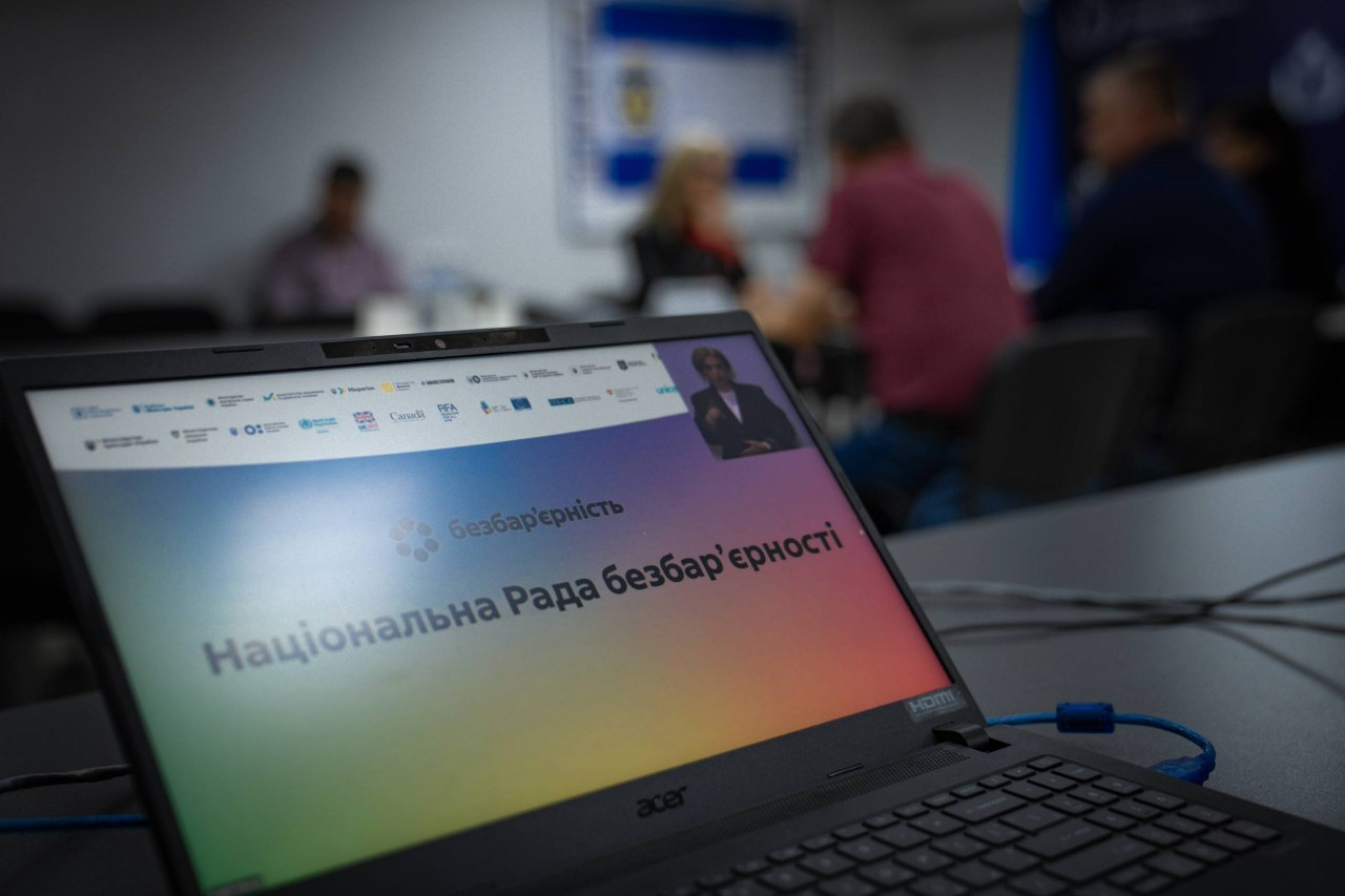 Екран чорного ноутбука на передньому плані відображає трансляцію засідання Національної Ради безбар’єрності з яскравою градієнтною заставкою, круглим логотипом і білим написом «Національна Рада безбар’єрності». У правому верхньому кутку вікна відеотрансляції в онлайн-режимі виступає перша леді Олена Зеленська, а на верхній панелі слайда розміщено логотипи міжнародних організацій-партнерів. На розмитому задньому плані видно залу засідань, де за робочим столом сидіння займають директорка Департаменту здоров’я Херсонської ОДА Тетяна Карчевич, директор Департаменту розвитку економіки Херсонської ОДА Артем Мішин, начальник управління фізичної культури, молоді та спорту Херсонської ОДА Сергій Яворський та інші учасники Національної Ради безбар’єрності від Херсонщини, що ілюструє участь посадовців у всеукраїнському обговоренні, за підсумками якого область посіла четверте місце в загальноукраїнському рейтингу безбар'єрності.