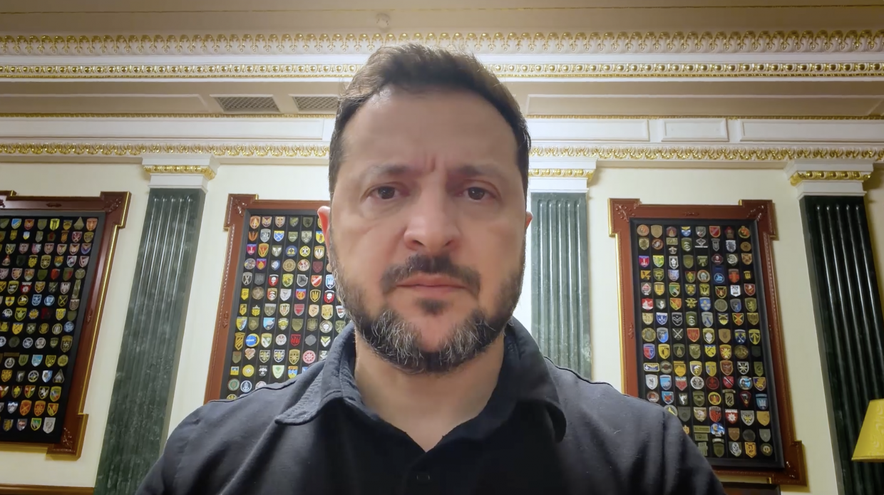Президент України Володимир Зеленський у чорній сорочці показаний крупним планом під час щоденного вечірнього відеозвернення до українців у робочому кабінеті Офісу Президента. Глава держави дивиться прямо в камеру на тлі класичного інтер'єру з білими стінами, оздобленими витонченою золотистою ліпниною, та декоративними колонами. На стінах за його спиною симетрично розміщено великі темні дерев'яні стенди з численними різнокольоровими шевронами та емблемами бойових бригад Збройних Сил України, а сам кадр супроводжує офіційну заяву про те, що найближчими днями очікується черговий транш фінансової бюджетної підтримки від Брюсселя, ухвалення рішень Бельгією щодо постачання додаткових літаків F-16 та посилення українських далекобійних завдань.