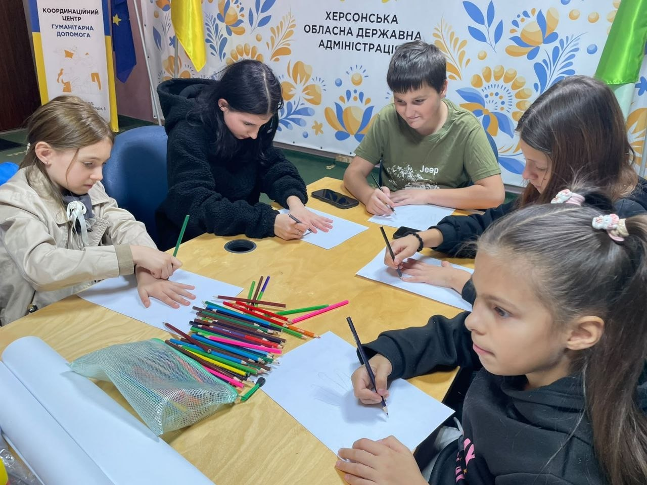 Діти захоплено малюють за великим дерев'яним столом у приміщенні Координаційного центру «Вільні разом» в Ужгороді. На передньому плані по центру розсипано купу різнокольорових олівців та лежить прозорий пенал, а хлопчик та дівчата різного віку зосереджено обводять свої долоні та створюють малюнки на білих аркушах паперу. Зустріч проходить на тлі великого світлого банера з патріотичними синьо-жовтими квітковими орнаментами та написом «ХЕРСОНСЬКА ОБЛАСНА ДЕРЖАВНА АДМІНІСТРАЦІЯ», поруч із яким видно державні прапори та жовтий штендер «Координаційний центр гуманітарна допомога», що ілюструє роботу дитячого простору центру, який протягом тижня приймав юних відвідувачів із родин переселенців з Херсонщини.