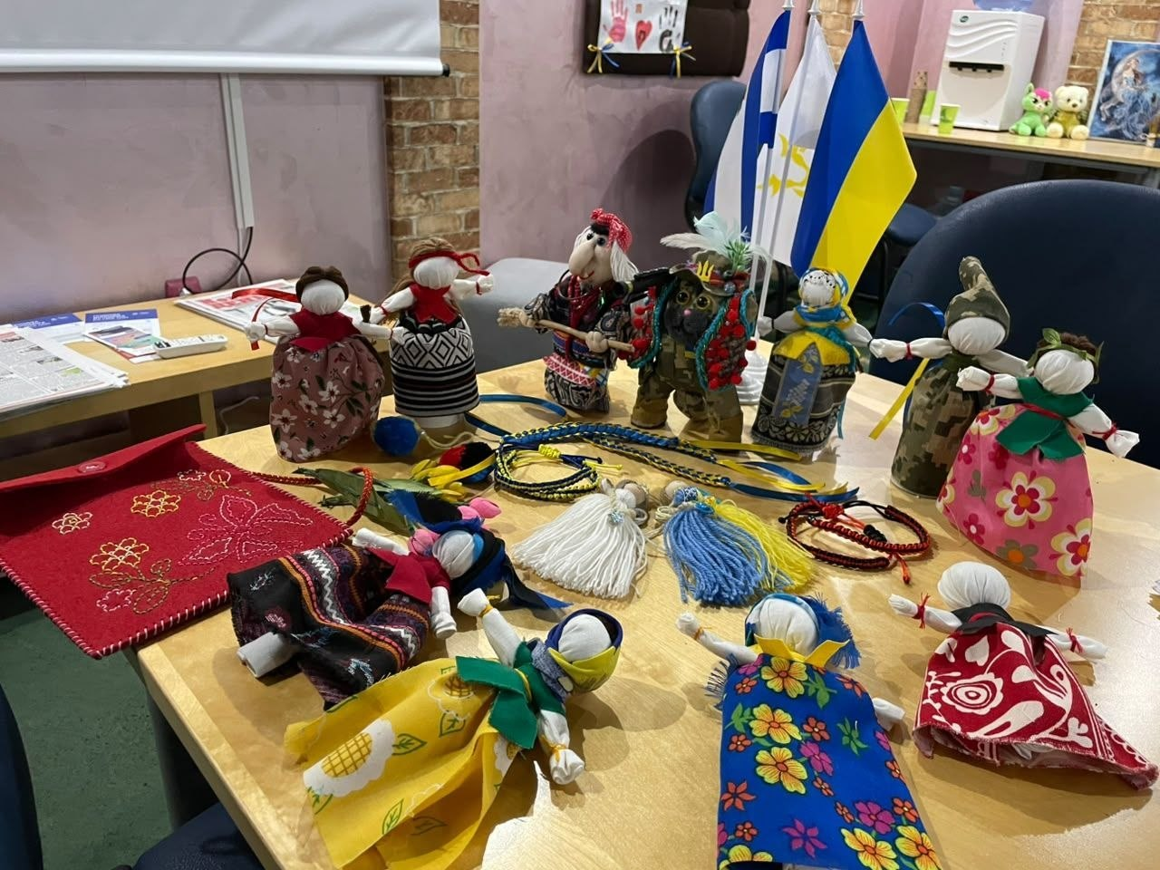 Різноманітні традиційні українські ляльки-мотанки та патріотичні обереги з ниток розкладені на великому дерев'яному столі за результатами проведення творчої майстерки «Моя мотанка, мій оберіг» в Ужгороді. На виставковій поверхні представлені вироби ручної роботи в автентичному вбранні, яскравих сукнях із квітковими та геометричними орнаментами, а також фігурки у військовому камуфляжі. На задньому плані по центру встановлено настільні Державні Прапори України та міста Херсона, що ілюструє культурно-освітній захід для психосоціальної підтримки переселенців з Херсонщини на базі Координаційного центру «Вільні разом».