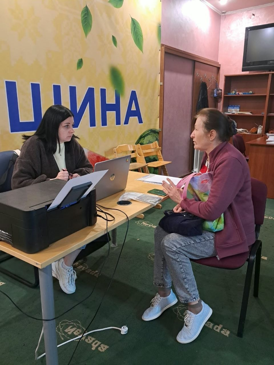 Юрисконсульт БФ «Право на захист» у коричневій кофті сидить за робочим столом перед відкритим ноутбуком HP та надає індивідуальну правову консультацію літній жінці в приміщенні Координаційного центру «Вільні разом» в Ужгороді. Фахівчиня уважно слухає відвідувачку, тримаючи в руці ручку поруч із настільним чорним принтером, а навпроти неї на стільці сидить жінка у бордовій куртці та сірих джинсах, яка жестикулює під час розмови. Зустріч проходить на тлі великого світлого банера з частиною напису «...ЩИНА» та зображенням зеленого листя, а сам кадр відображає процес надання правової підтримки переселенцям з Херсонщини в евакуації, у межах якого юристи благодійного фонду проконсультували 7 учасників.