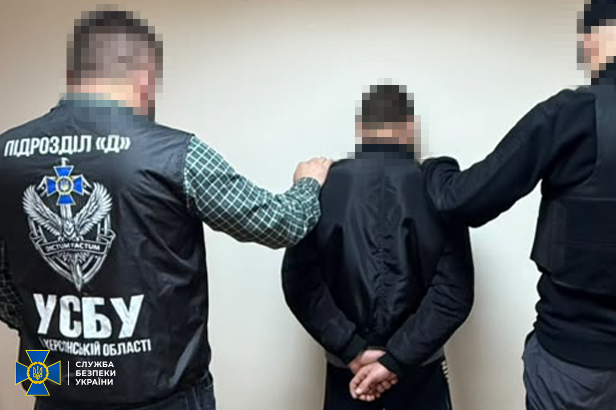 Двоє оперативників спецслужби тримають затриманого колаборанта зі згорнутими за спиною руками на тлі світлої стіни кабінету під час спільної операції правоохоронних органів. Ліворуч спиною до камери стоїть співробітник у фірмовому чорному тактичному жилеті з білими написами «ПІДРОЗДІЛ 