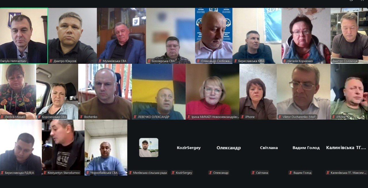 Екран монітора відображає велику сітку вікон онлайн-зв'язку програми Zoom під час робочої наради під головуванням голови Комітету Верховної Ради України з питань фінансів, податкової та митної політики Данила Гетманцева. Численні учасники зустрічі зафіксовані у своїх віконцях під час обговорення, причому у лівому верхньому кутку транслюється сам Данило Гетманцев (підпис «Danylo Hetmantsev»), поруч із ним — директор Департаменту розвитку сільського господарства та зрошення Херсонської ОДА Дмитро Юнусов, а також перший заступник начальника Херсонської ОВА Дмитро Бутрій. Серед інших вікон присутні народні депутати, представники міністерств, керівники військових адміністрацій: Музиківської СВА, Білозерської СВА, Бериславської МВА, Борозенської СВА, Чорнобаївської СВА, Милівської сільської ради, Калинівської ТГ, виконавчий директор Асоціації міст України Олександр Слобожан та інші. Кадр ілюструє процес дистанційного обговорення законодавчих змін щодо зниження ставок мінімального податкового зобов’язання для аграріїв Херсонщини, які обробляють землю в зонах бойових дій.