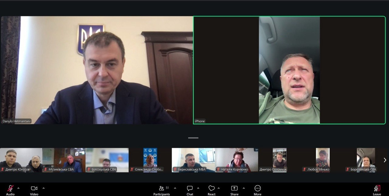 Екран монітора відображає вікно онлайн-зв'язку програми Zoom під час робочої наради, де у двох великих центральних вікнах транслюються голова Комітету Верховної Ради України Данило Гетманцев та перший заступник начальника Херсонської ОВА Дмитро Бутрій. Ліворуч на передньому плані Данило Гетманцев (підпис «Danylo Hetmantsev») зафіксований у кабінеті на тлі Державного Герба України, а праворуч у великому вікні (підпис «iPhone») підключений Дмитро Бутрій у військовій футболці кольору хакі, який виступає безпосередньо з автомобіля. На нижній горизонтальній панелі інтерфейсу розміщено мініатюри інших вікон учасників, серед яких видно директора Департаменту розвитку сільського господарства Дмитра Юнусова, представників Музиківської СВА, Білозерської СВА, Бериславської МВА, Борозенської СВА, виконавчого директора Асоціації міст України Олександра Слобожана та інших, що ілюструє спільне обговорення зниження податкового зобов’язання для аграріїв Херсонщини у зоні бойових дій.