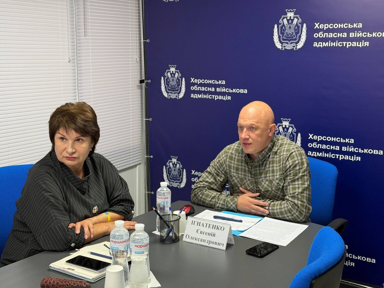 Заступник начальника Херсонської ОВА Євгеній Ігнатенко та заступниця начальника Херсонської МВА Наталія Чорненька сидять за робочим столом у кабінеті під час онлайн-засідання Міністерства соціальної політики України щодо підготовки області до опалювального сезону. Праворуч у картованій сорочці сидіння займає Євгеній Ігнатенко, розклавши перед собою робочі папери з ручкою. Ліворуч у темній смугастій блузі сидить Наталія Чорненька, яка уважно слухає доповіді колег. Засідання проходить на тлі великого синього фірмового банера з офіційними гербами та повторюваними написами «Херсонська обласна військова адміністрація», а сам кадр відображає процес координації виплат та забезпечення жителів прифронтових громад твердим паливом.