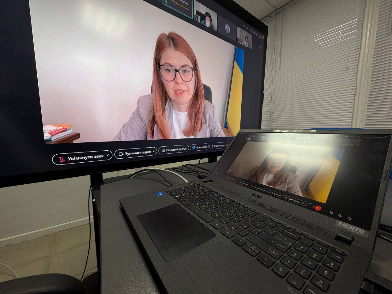Великий екран монітора та підключений до нього чорний ноутбук Acer відображають вікно онлайн-зв'язку під час засідання Міністерства соціальної політики України щодо підготовки Херсонської області до опалювального сезону. На великому дисплеї по центру крупним планом транслюється виступ заступниці Міністра соціальної політики України Мар'яни Токар в окулярах та сірому жакеті, яка виступає на тлі Державного Прапора України, а у верхній панелі інтерфейсу та на екрані ноутбука меншими віконцями відображаються інші учасники дистанційної зустрічі. Кадр знято збоку і зблизька, що ілюструє процес цифрової координації соціальних виплат та забезпечення паливом жителів прифронтових громад.