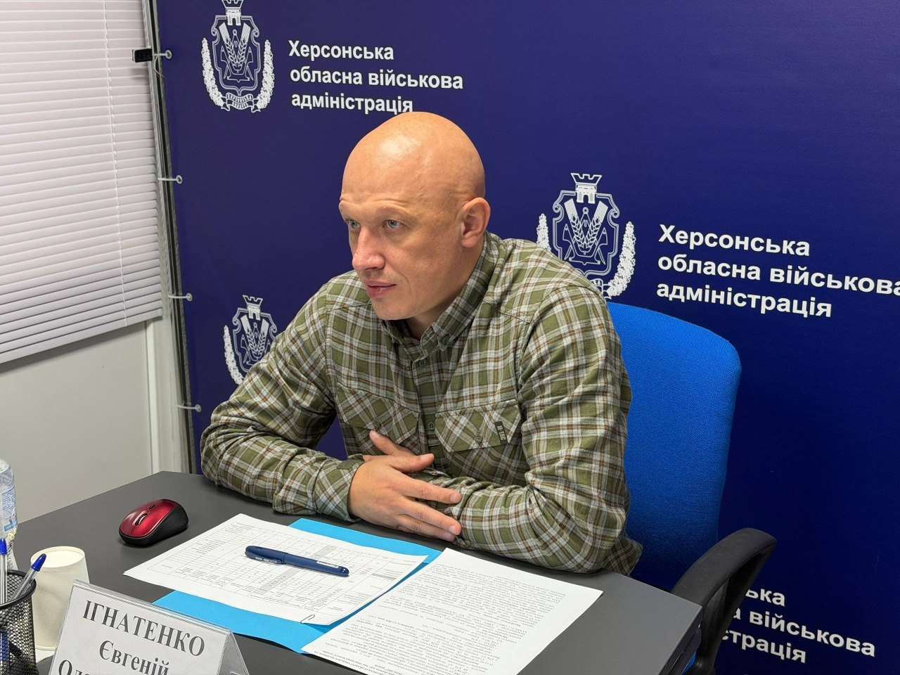 Заступник начальника Херсонської ОВА Євгеній Ігнатенко бере участь в онлайн-засіданні Міністерства соціальної політики України щодо підготовки області до опалювального сезону. Посадовець у картованій сорочці сидить по центру кадру за робочим столом, склавши руки перед собою, та уважно слухає доповіді колег. На столі перед ним розкладено друковані бланки документів, папери та ручку, а ліворуч на передньому плані частково видно комп'ютерну мишу та край іменної таблички. Робоче місце облаштоване на тлі великого синього фірмового банера з офіційними гербами та повторюваними написами «Херсонська обласна військова адміністрація», а сам кадр відображає процес координації забезпечення жителів прифронтових громад твердим паливом.