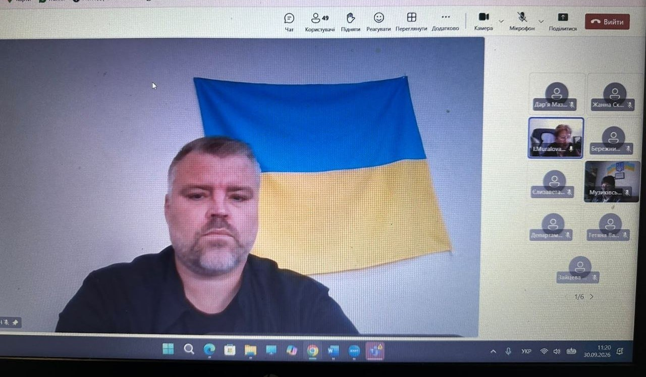 Екран монітора відображає вікно онлайн-зв'язку під час навчального семінару для представників Херсонщини, де у великому центральному вікні крупним планом транслюється директор Департаменту розвитку економіки Херсонської ОДА Артем Мішин. Посадовець слухає колег на тлі Державного Прапора України, закріпленого на світлій стіні. У правій частині інтерфейсу відображається вертикальна панель із мініатюрними іконками інших учасників онлайн-зустрічі. Кадр ілюструє координацію та проведення дистанційного заходу з питань реалізації державної регуляторної політики та підтримки бізнес-середовища в регіоні.