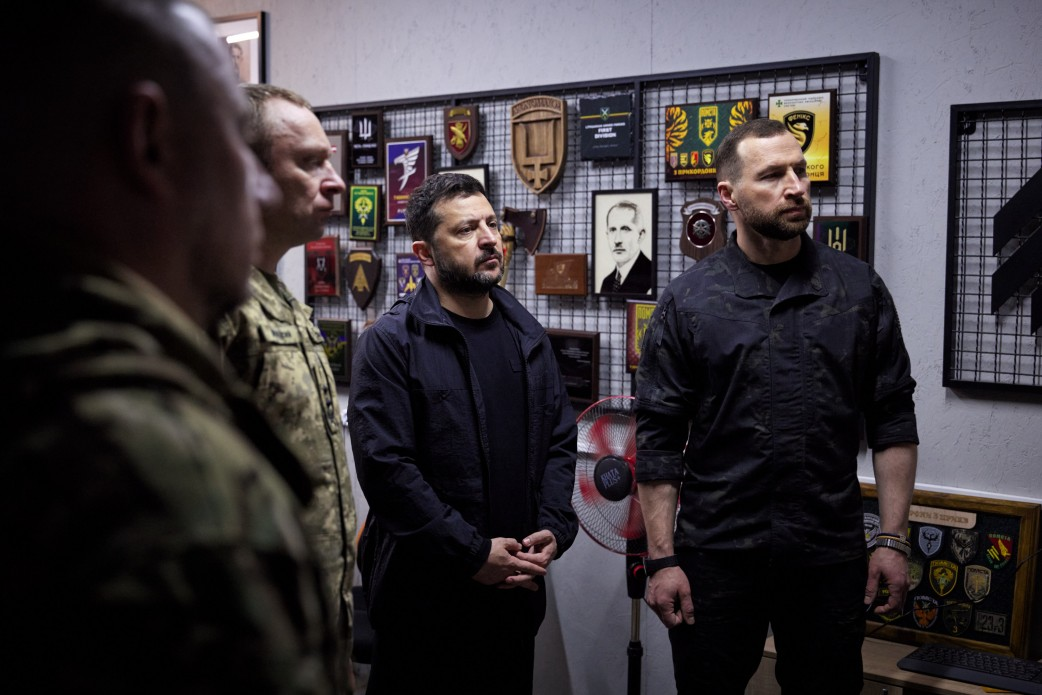 Президент України Володимир Зеленський та Міністр оборони України Євгеній Хмара під час робочої поїздки на Лиманський напрямок та зустрічі з військовослужбовцями у прифронтовому пункті управління з нагоди Дня захисників і захисниць України. По центру кадру стоїть Володимир Зеленський у чорній футболці та темній тактичній куртці, склавши руки перед собою та уважно слухаючи доповіді командирів. Праворуч від нього показаний Міністр оборони Євгеній Хмара у чорній військовій сорочці із загорнутими рукавами. Ліворуч у профіль та напівпрофіль стоять українські захисники у камуфляжній формі. Зустріч проходить на тлі світлої стіни, яку прикрашено металевою сіткою-стендом із численними військовими шевронами, пам'ятними відзнаками та дерев'яними коїнами підрозділів, що ілюструє перебування лідерів безпосередньо на передовій у 1681-шу добу повномасштабної війни.