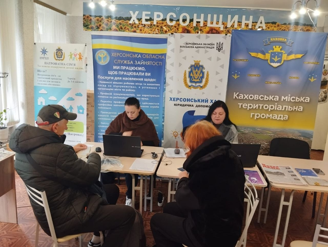 Фахівці з надання правової допомоги та представники державних служб консультують внутрішньо переміщених осіб із Херсонщини за двома зсунутими разом столами в приміщенні хабу. Ліворуч за столом перед відкритим ноутбуком сидить спеціалістка, яка спілкується з літнім чоловіком у чорній куртці та бейсболці. Праворуч за сусіднім столом інша фахівчиня роз'яснює юридичні питання відвідувачці з рудим волоссям, яка сидить на стільці спиною до камери. Зустріч проходить на тлі великого банера з написом «ХЕРСОНЩИНА» та зображенням соняшників, синього банера Каховської міської територіальної громади, ролапів Херсонської обласної служби зайнятості та Херсонського хабу, а сам кадр відображає процес надання комплексної безкоштовної правової підтримки переселенцям в евакуації у Києві.
