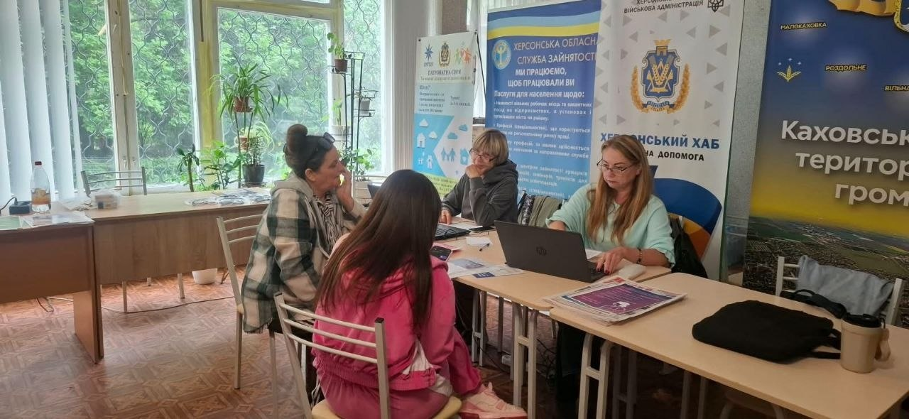 Дві фахівчині гуманітарного хабу за довгим робочим столом надають індивідуальні консультації внутрішньо переміщеним особам із Херсонщини в межах роботи координаційного центру. Праворуч перед відкритим ноутбуком сидить спеціалістка в окулярах та світло-зеленій блузі, яка веде діалог із дівчиною у рожевому спортивному костюмі, що сидить навпроти спиною до камери. Поруч за цим же столом інша фахівчиня у сірій кофті працює за своїм ноутбуком та консультує жінку у картованій сорочці. Зустріч проходить у світлій кімнаті з великими вікнами та кімнатними рослинами на тлі ролапів Херсонської обласної служби зайнятості, Херсонського хабу та інформаційного банера Каховської міської територіальної громади, що ілюструє процес надання комплексної правової та психосоціальної підтримки переселенцям в евакуації у Києві.