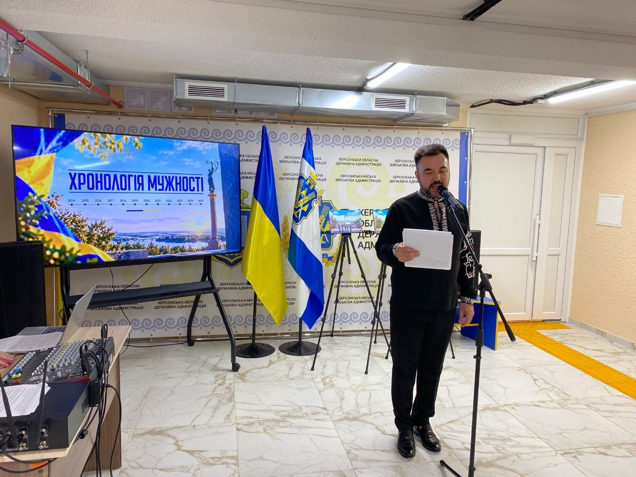 Заступник директора Департаменту реалізації гуманітарної політики Херсонської ОДА Андрій Ярьоменко у вишиванці зачитує промову перед мікрофоном під час урочистого заходу «Під покровом мужності» до Дня захисників і захисниць України. Чоловік у чорній сорочці з білим орнаментом і чорних штанях стоїть праворуч біля мікрофонної стійки, тримаючи в руках аркуш паперу. Зустріч проходить в облаштованому залі на тлі Державного Прапора України та прапора Херсонської області. Ліворуч на передньому плані розташовано апаратуру звукорежисера, а за нею встановлено великий мультимедійний екран, на якому демонструється презентація книги пам'яті «ХРОНОЛОГІЯ МУЖНОСТІ» із зображенням херсонського монумента на площі Свободи. Задній план оформлено великим білим банером із повторюваними написами «Херсонська обласна державна адміністрація» та «Херсонська міська військова адміністрація».