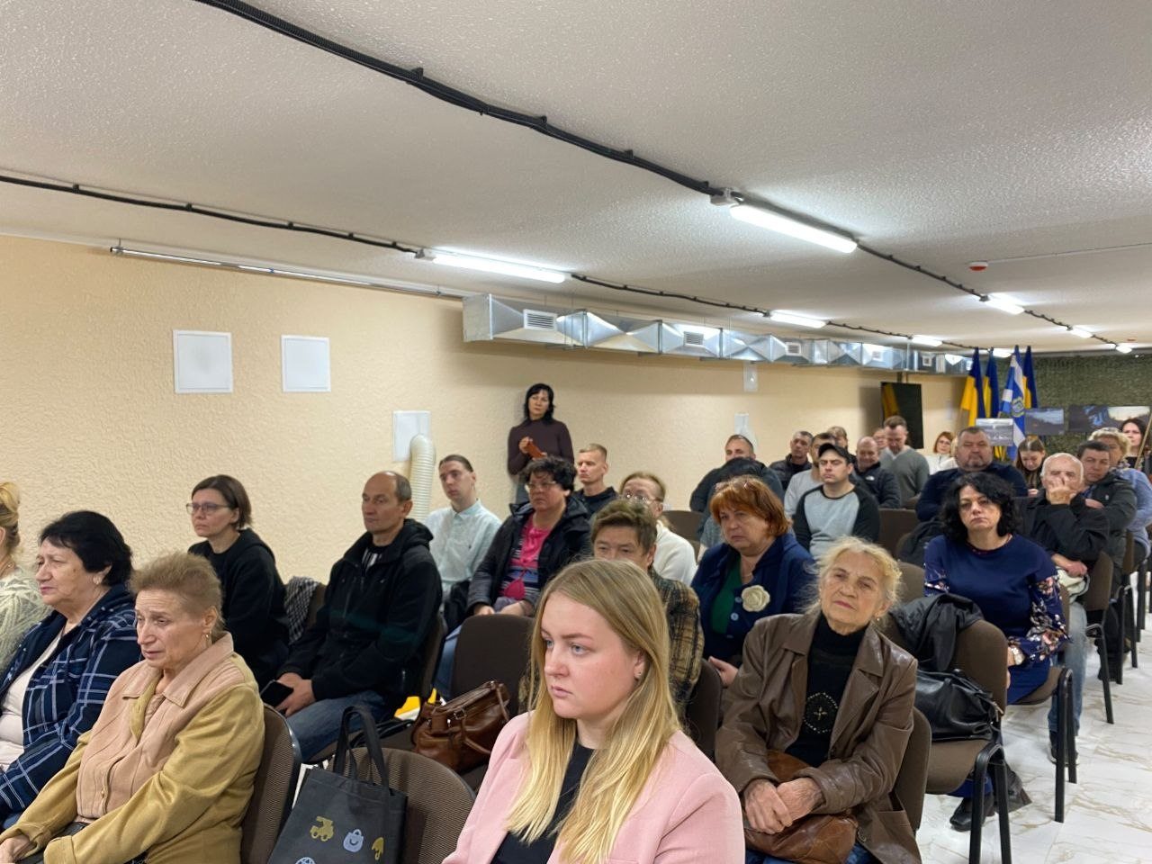Присутні на заході «Під покровом мужності» сидять у конференц-залі в Херсоні, уважно слухаючи виступи під час презентації літопису «Хронологія мужності». Захід організовано Департаментом реалізації гуманітарної політики Херсонської ОВА у співпраці з Управлінням культури Херсонської МВА. На передньому плані праворуч виділяється молода жінка зі світлим волоссям у рожевому жакеті, а поруч із нею — літня жінка в коричневій шкіряній куртці. У наступних рядах видно інших мешканців Херсонщини та родичів полеглих воїнів. На задньому плані праворуч видніються державні прапори України та прапор Херсонської області.