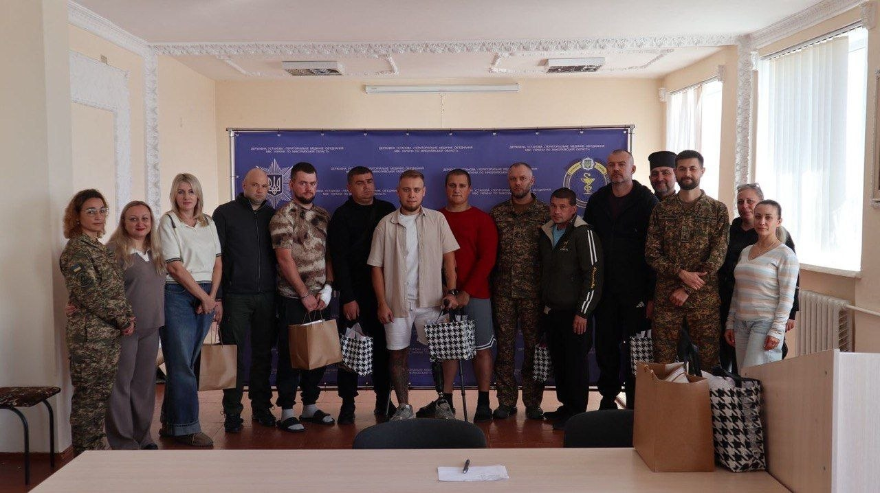 Групове фото у світлій кімнаті: представники Херсонської ОДА, Національної гвардії України та поранені українські військовослужбовці, які проходять лікування й реабілітацію. У центрі стоять чоловіки-військові (частина з них у цивільному одязі, шортах та футболках, дехто спирається на милиці або має видимі ознаки травм та протезування після поранень). Поруч із ними стоять військовослужбовці НГУ в камуфляжній формі. Крайньою праворуч у світлій кофті в смужку та блакитних джинсах стоїть начальниця управління у справах ветеранів Херсонської ОДА Дар’я Мазур. На передньому плані видно довгий стіл, на якому стоять великі пакунки з подарунками для захисників. На задньому плані розміщено великий синій банер із державною символікою України.