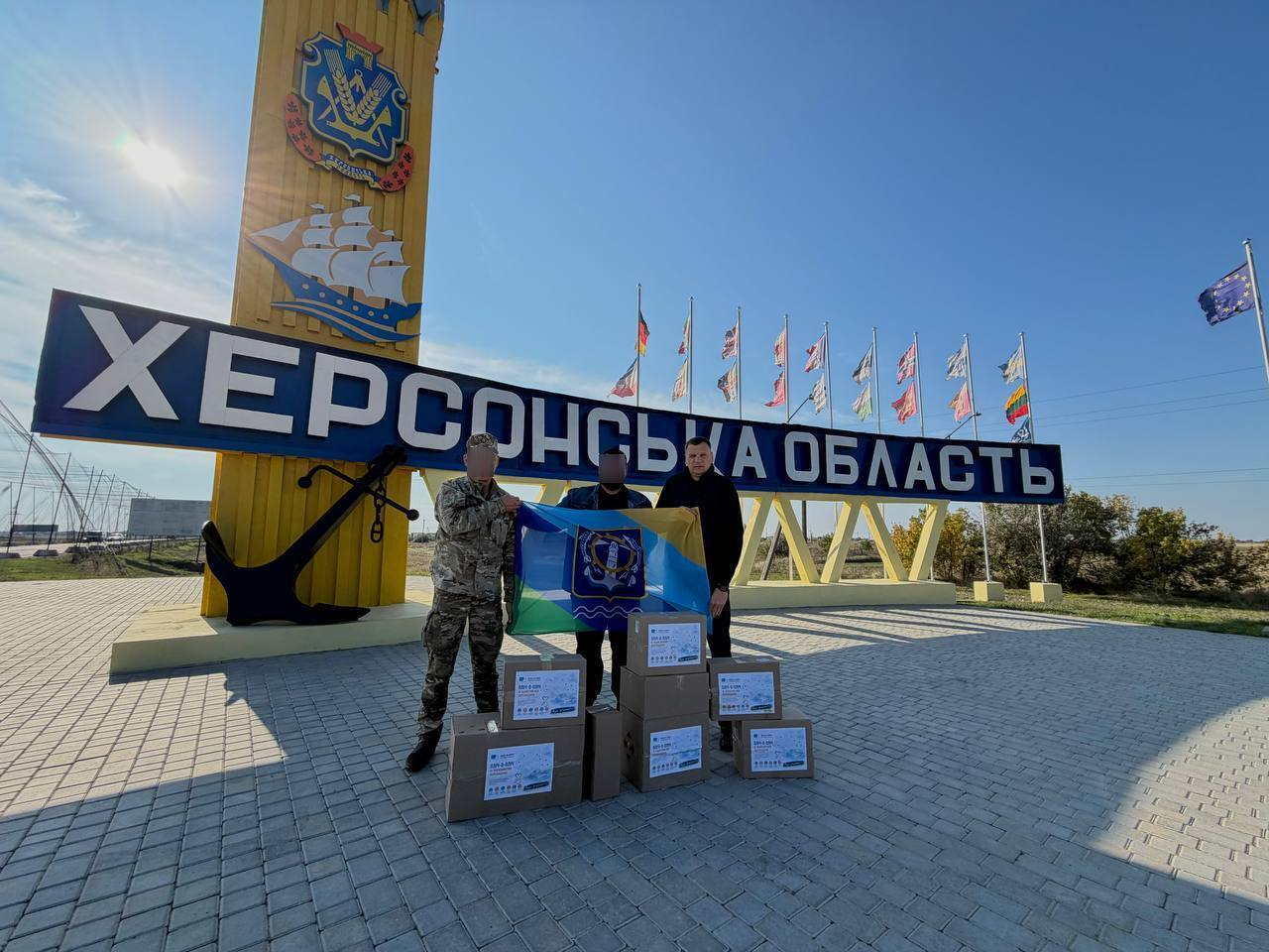 Групове фото на тлі стели «Херсонська область»: троє чоловіків стоять біля великого жовтого в'їзного знака з гербом Херсонщини та зображенням вітрильника. Ліворуч стоїть військовий у камуфляжній формі, а по центру — чоловік у цивільному одязі, обличчя обох приховані з міркувань безпеки. Праворуч стоїть начальник Генічеської МВА Євгеній Родіонов у чорній куртці, який дивиться в кадр. Разом вони тримають розгорнутий бойовий прапор 79-го прикордонного загону ДПСУ. Перед ними на бруківці складено кілька картонних коробок із гуманітарною допомогою, на кожній з яких наклеєно біло-блакитні інформаційні аркуші. На задньому плані, за написом «ХЕРСОНСЬКА ОБЛАСТЬ», височіє ряд флагштоків із прапорами, а праворуч на тлі чистого синього неба майорить прапор Європейського Союзу.