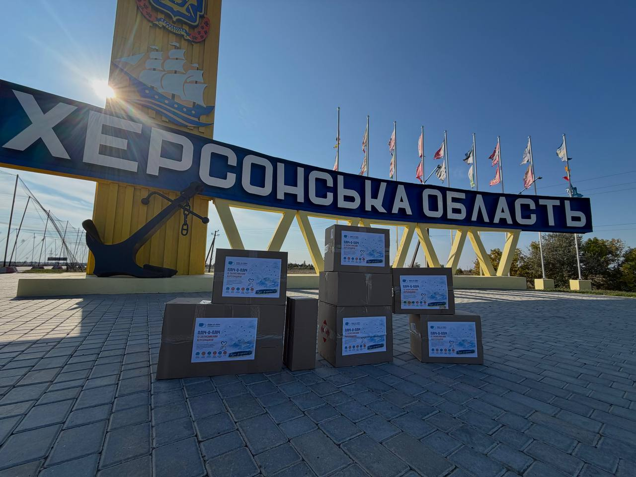 На передньому плані кадру на бруківці перед стелою «Херсонська область» великим планом викладено вісім картонних коробок різного розміру з гуманітарною допомогою, які складені у кілька невеликих груп. На лицьовому боці кожної коробки наклеєно біло-блакитний інформаційний аркуш із логотипом благодійної акції «Пліч-о-пліч із захисниками Херсонщини». За коробками височіє жовтий в'їзний знак із гербом Херсонщини та вітрильником, під яким закріплено масивний темний якір і встановлено великий синій напис «ХЕРСОНСЬКА ОБЛАСТЬ». На задньому плані за конструкцією знака під ясним небом вишикувано ряд флагштоків із прапорами, а яскраве сонце освітлює стелу ліворуч, створюючи невеликі відблиски.