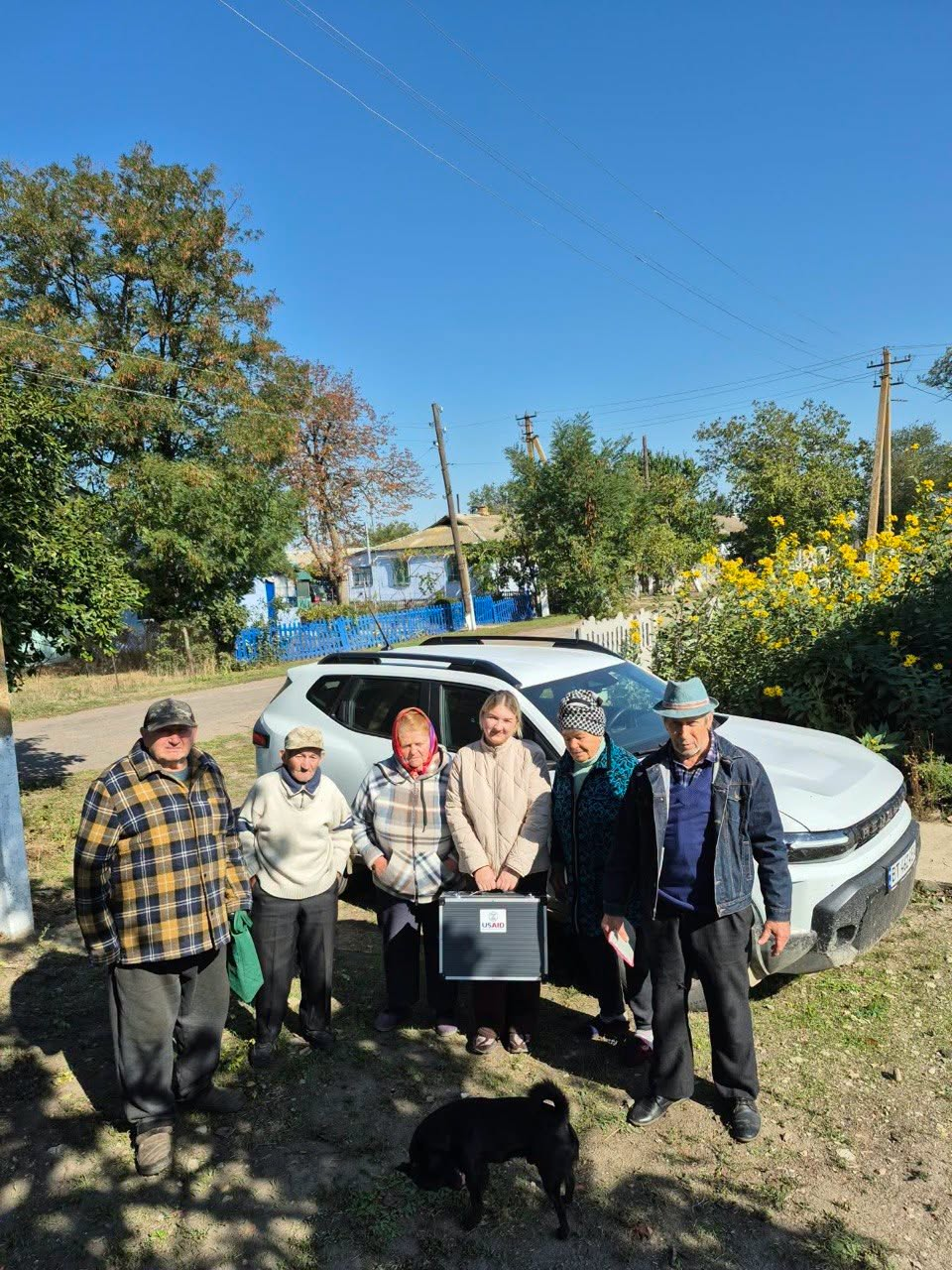 Групове фото на вулиці у сонячний день: шестеро людей стоять перед білим кросовером на тлі сільського пейзажу з деревами, будинками та синім парканом. У центрі групи стоїть молода дівчина, працівниця ЦНАПу, яка тримає в руках закриту металеву мобільну валізу з логотипом USAID. Її оточують п'ятеро місцевих жителів похилого віку — троє чоловіків та дві жінки у теплому повсякденному одязі, куртках і головних уборах. На передньому плані на землі перед людьми стоїть невеликий чорний собака.