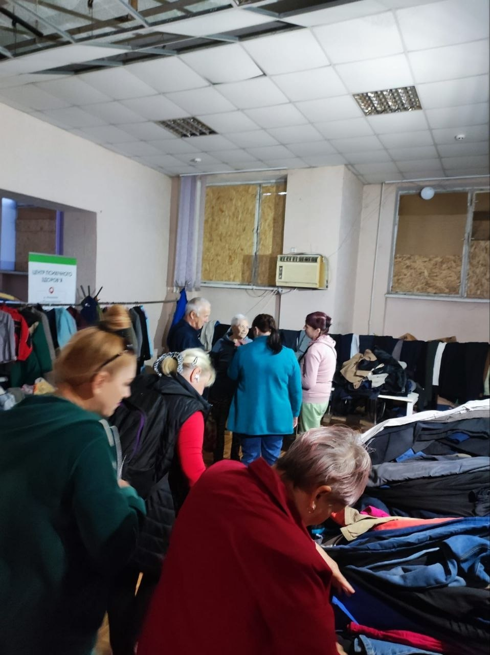 У Координаційному центрі «Вільні разом» у Миколаєві люди обирають вживаний одяг, наданий Фондом громади міста Херсон «Захист». На передньому плані жінки схилилися над великими стопками різнокольорових речей, перебираючи їх. Трохи далі в глибині кімнати стоїть група людей, які спілкуються між собою. Ліворуч облаштовано вішалки із верхнім одягом, біля яких встановлено інформаційний стенд із написом «Центр психічного здоров'я». Приміщення освітлюється стельовими світильниками, вікна забиті плитами ОСБ, а під стелею видно елементи підвісної конструкції.