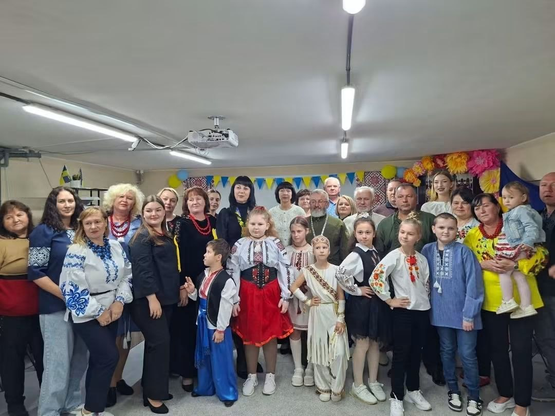 Велика група дітей та дорослих позує для спільного фото в приміщенні Центру соціальної підтримки дітей та сімей з нагоди Покрови Пресвятої Богородиці, Дня українського козацтва та Дня захисників і захисниць України. У самому центрі кадру стоїть начальниця Служби у справах дітей Херсонської ОДА Тетяна Лушнікова, вбрана в чорний костюм, а праворуч від неї — Алла Малицька, заступниця Херсонського міського голови з питань діяльності виконавчих органів ради. Також на світлині присутні працівники закладу та вихованці центру, частина з яких одягнена в українські національні костюми, вишиванки та шаровари. Кімната прикрашена синьо-жовтими прапорцями, кульками та квітами, а на стелі ввімкнено сучасне лінійне освітлення та встановлено проектор.
