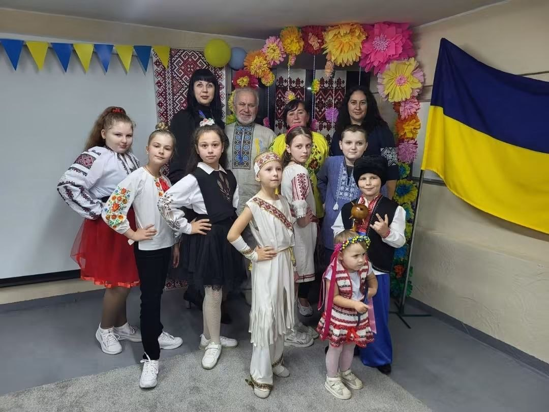 Група вихованців та працівників Центру соціальної підтримки дітей та сімей разом із начальницею Служби у справах дітей Херсонської ОДА Тетяною Лушніковою позують для фотографії під час заходу до Дня захисників і захисниць України. На передньому плані стоять діти у різноманітних вишиванках, шароварах та традиційному українському вбранні, а один хлопчик тримає в руках сувенірний козацький куманець (посуд). На задньому плані поруч із дорослими стоїть літній чоловік у вишитій сорочці. Локація святково прикрашена аркою з великих паперових квітів, жовто-блакитними прапорцями-гірляндами, а праворуч на стіні закріплено великий державний прапор України.