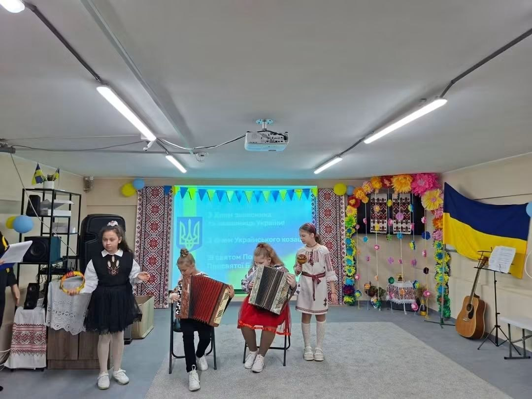 Чотири вихованки Центру соціальної підтримки дітей та сімей виступають на сцені під час творчих активностей до Дня захисників і захисниць України. Дві дівчинки сидять на стільцях у центрі та грають на маленьких акордеонах (гармошках), тоді як дві інші дівчинки стоять обабіч них у традиційних українських сукнях та вишиванках, тримаючи в руках народні шумові інструменти — бубон та маракас. На задньому плані працює великий проекційний екран із тризубом та привітальним текстом до Дня козацтва та Покрови, оточений банерами з вишитим орнаментом та квітковою аркою. Праворуч на сцені біля великого державного прапора України стоїть акустична гітара на підставці та пюпітр із нотами.