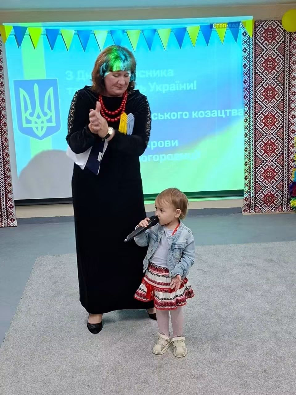 Маленька дівчинка, вихованка Центру соціальної підтримки дітей та сімей, вбрана у джинсову куртку та спідницю з українським народним орнаментом, виступає на сцені та тримає у руці великий мікрофон. Поруч із нею стоїть доросла жінка у довгій чорній сукні з червоним намистом, яка підтримує маленьку артистку та плескає в долоні. Цей зворушливий момент зафіксовано під час святкового заходу до Дня захисників і захисниць України, Покрови та Дня козацтва. На задньому плані працює великий мультимедійний екран із зображенням Герба України (Тризуба) та привітальними написами, а обабіч нього встановлені декоративні банери з традиційною українською геоmetric вишивкою.