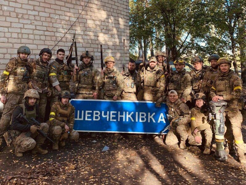 Група українських військовослужбовців у повному бойовому екіпіруванні, камуфляжній формі та шоломах позує для фотографії під час Херсонської наступальної операції у жовтні 2022 року, в ході якої Сили оборони звільнили 41 населений пункт Бериславського району. У центрі кадру бійці тримають великий синій дорожній вказівник із білим написом «ШЕВЧЕНКІВКА», що символізує повернення свободи та українського прапора до окупованих ворогом селищ. Позаду військових стоїть автомобіль та цегляна будівля, а самі захисники тримають у руках автоматичну зброю та переносний протитанковий комплекс.