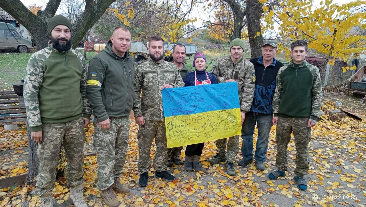 Група українських військових у камуфляжній формі та місцеві жителі Бериславського району разом тримають великий синьо-жовтий прапор України, вкритий підписами та побажаннями, на тлі осінніх дерев із жовтим листям. Знімок зроблено наприкінці жовтня 2022 року під час успішної Херсонської наступальної операції, коли Сили оборони звільнили 41 населений пункт правобережжя, повернувши свободу місцевим родинам після понад пів року російської окупації. На подвір'ї поруч із цивільними та військовими видно сільську техніку.
