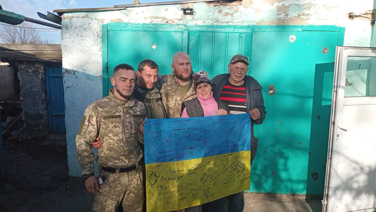 Троє українських військових у камуфляжній формі та двоє цивільних місцевих жителів позують разом на тлі яскраво-бірюзових воріт гаража під час Херсонської наступальної операції у жовтні 2022 року. Вони посміхаються та тримають перед собою великий розгорнутий прапор України, густо вкритий патріотичними підписами, побажаннями та назвами міст від руки. Цей кадр символізує звільнення Бериславського району правобережжя Херсонщини від піврічної російської окупації та довгоочікуване повернення свободи для місцевих родин, які зустрічали Сили оборони як справжніх Героїв. На стіні будівлі видно сліди від уламків, а у правому нижньому кутку фото присутній напівпрозорий часовий штамп.