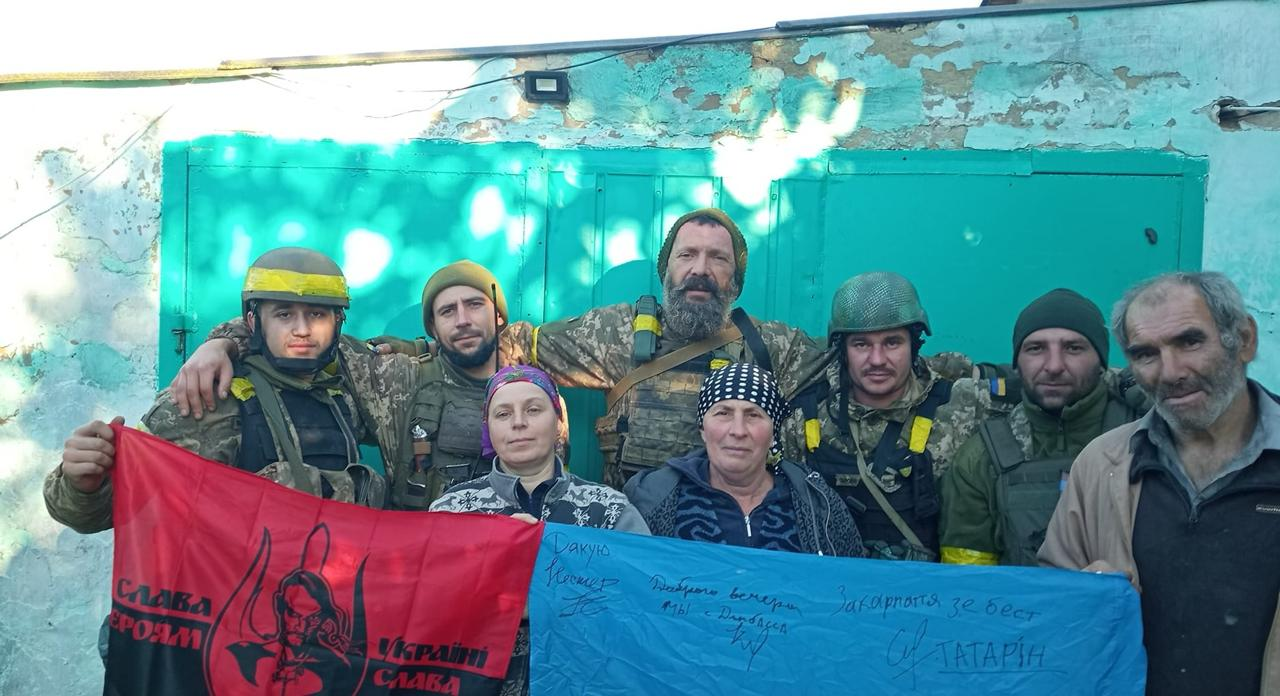 П'ятеро українських військовослужбовців у повному екіпіруванні, бронежилетах та шоломах позують в обіймах із трьома цивільними місцевими жителями на тлі бірюзової стіни будівлі під час Херсонської наступальної операції у жовтні 2022 року. У руках вони тримають два прапори: червоно-чорний стяг із написом «Слава Героям – Україні Слава» та малюнком козака, а також синю частину полотна, вкриту рукописними підписами та фразами, серед яких видно «Дякую» та «Закарпаття зе бест». Фотографія передає щиру радість та полегшення людей Бериславського району, звільнених Силами оборони після понад пів року жорсткої російської окупації правобережжя Херсонщини.