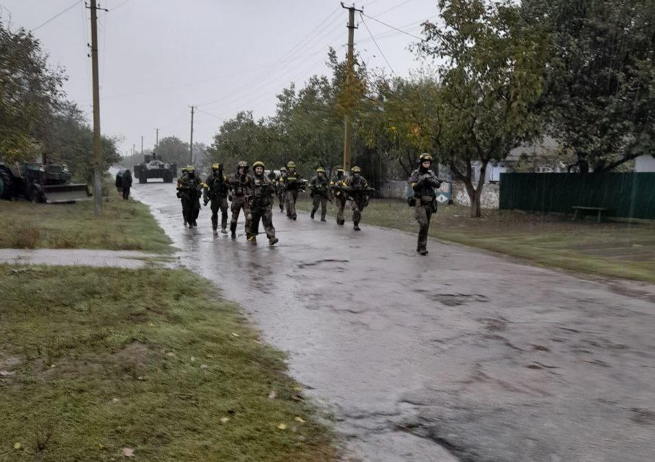 Підрозділ українських військовослужбовців у повному бойовому екіпіруванні та шоломах рухається пішим маршем по вологій асфальтованій дорозі через населений пункт під час Херсонської наступальної операції у жовтні 2022 року. Захисники йдуть зі зброєю в руках двома колонами вздовж узбіччя, а попереду них, випереджаючи групу, рухається один боєць. На задньому плані, в кінці вулиці, видно військову бронетехніку та силуети людей, а обабіч дороги розташовані одноповерхові сільські будинки, паркани, стовпи електропередач та зелені дерева. Кадр зроблений у похмуру дощову погоду та задокументував історичний момент просування Сил оборони, які звільнили 41 населений пункт Бериславського району правобережжя Херсонщини від тривалої російської окупації.