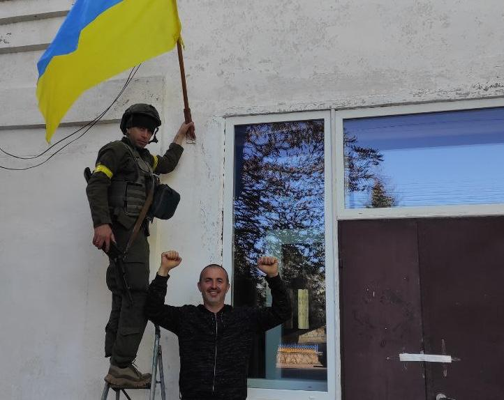 Український військовослужбовець у камуфляжній формі, бронежилеті та шоломі стоїть на металевій драбині біля стіни будівлі та встановлює синьо-жовтий прапор України. Поруч із ним, на землі, стоїть усміхнений цивільний чоловік, який радісно підняв угору зігнуті в ліктях руки із затиснутими кулаками, святкуючи звільнення. Цей момент зафіксовано під час успішної Херсонської наступальної операції у жовтні 2022 року, коли Сили оборони вибили ворога з 41 населеного пункту Бериславського району правобережжя, повернувши свободу місцевим жителям після понад пів року російської окупації. На задньому плані видно світлу стіну споруди, велике металопластикове вікно та темні вхідні двері.