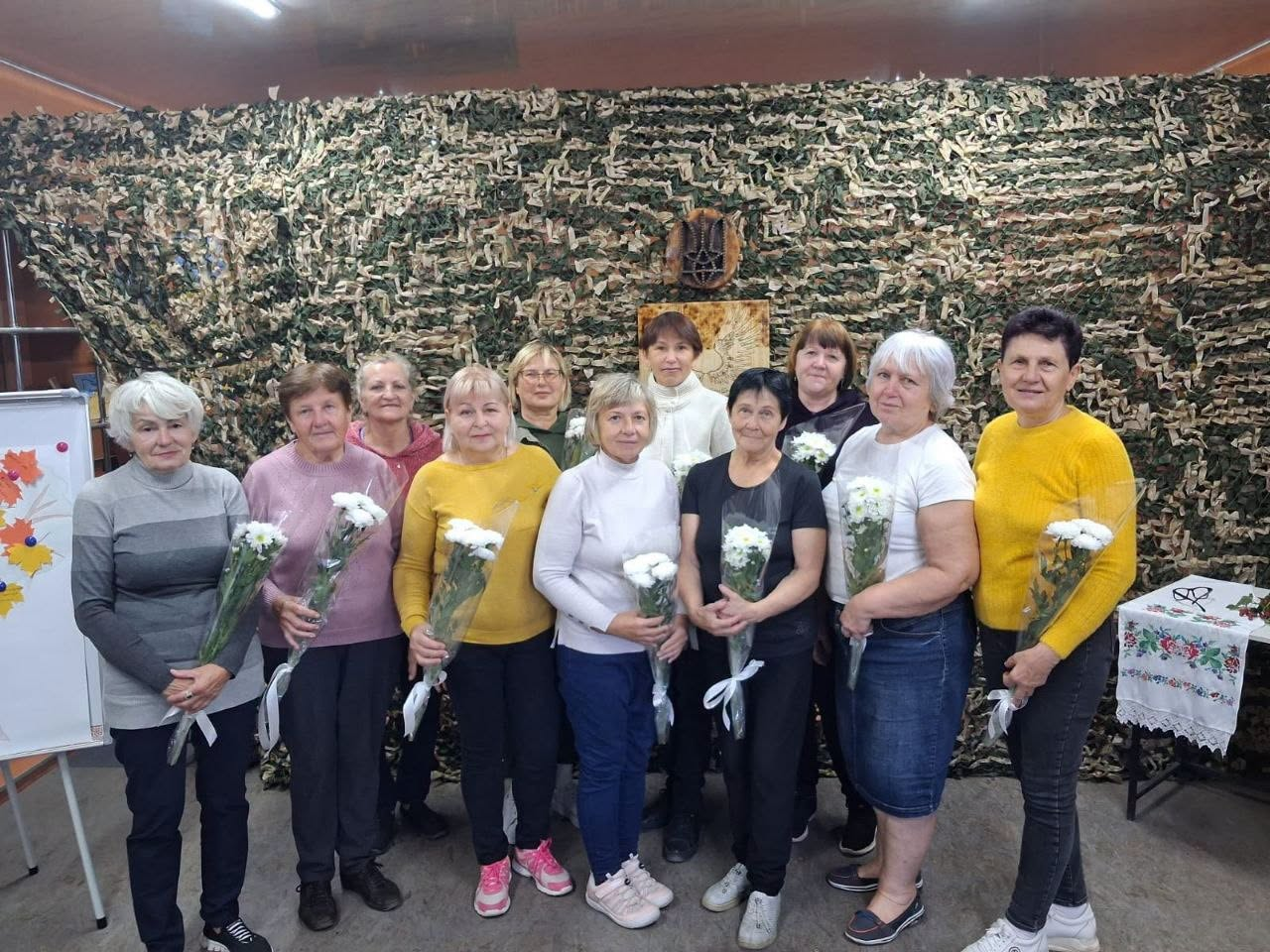 Група волонтерок Бериславської громади позує для спільного фото на тлі великої власноруч виготовленої маскувальної сітки зеленого та коричневого кольорів, яка призначена для захисту позицій і техніки воїнів на передовій. Кожна жінка тримає в руках букет білих хризантем, перев'язаний стрічкою. По центру над ними на сітці закріплений дерев'яний декоративний елемент із зображенням Герба України (Тризуба). Ліворуч у кадрі видно фліпчарт із дитячими малюнками осіннього листя, а праворуч — столик, накритий вишитим рушником. Світлина зафіксувала момент вдячності місцевим жителькам за системну підтримку Сил оборони та передачу чергової партії маскувального покриття.