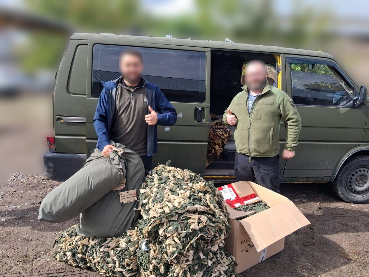Двоє чоловіків стоять біля військового мікроавтобуса кольору хакі із відчиненими боковими дверима та показують жест «великий палець угору». На землі перед ними лежить великий зелений волонтерський мішок із речами, оберемок виготовленої волонтерками Бериславської громади маскувальної сітки зеленого та бежевого кольорів, а також відкрита картонна коробка з іншою допомогою. Кадр зафіксував момент передачі чергової партії маскувального покриття воїнам на передовій для надійного укриття фронтових позицій, бліндажів та техніки від розвідувальних і ударних безпілотників ворога.