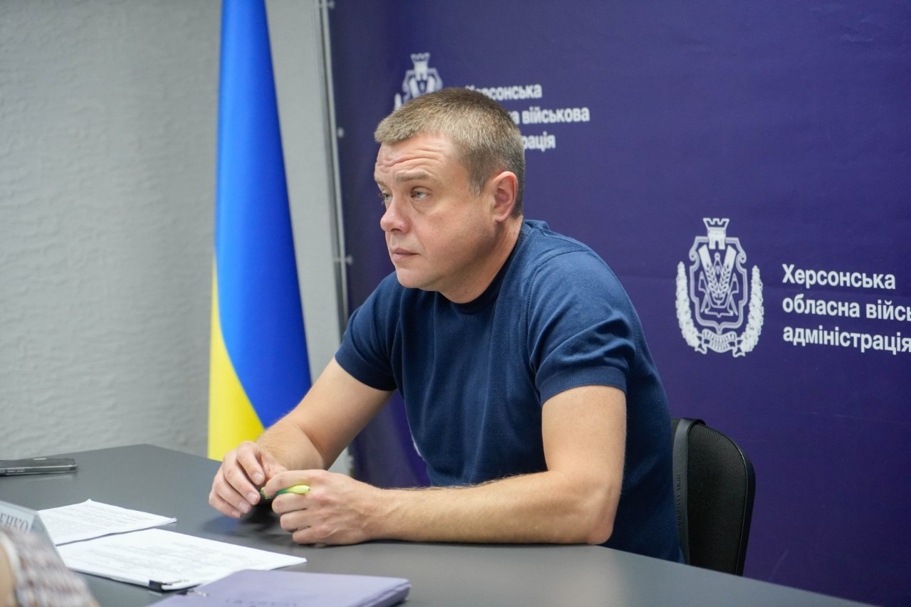 Заступник начальника Херсонської ОВА Антон Самойленко сидить за столом під час спільної наради щодо підготовки області до осінньо-зимового періоду 2026/2027 року. Посадовець вбраний у темно-синю футболку, тримає в руках ручку і зосереджено слухає учасників зустрічі. На столі перед ним лежать документи та блокнот. За ним, на задньому плані ліворуч, встановлено великий державний прапор України, а праворуч розташований фіолетовий банер із офіційним гербом та написом «Херсонська обласна військова адміністрація». Світлина ілюструє координацію планів із Міністерством відновлення для посилення енергетичної та інфраструктурної стійкості правобережжя до настання холодів.