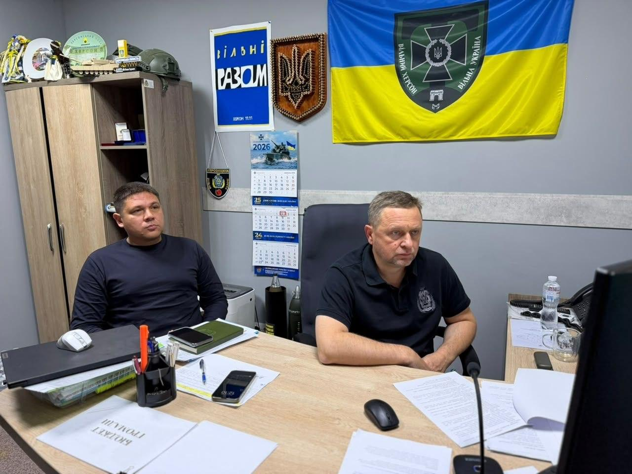 Перший заступник начальника Херсонської ОВА Дмитро Бутрій (праворуч) та директор Департаменту розвитку сільського господарства та зрошення Херсонської ОДА Дмитро Юнусов (ліворуч) сидять за робочим столом у кабінеті під час наради із заступником Міністра аграрної політики та продовольства України Віталієм Головнею щодо підтримки прифронтових аграріїв та вивезення врожаю з небезпечних територій області. Обидва посадовці вбрані у темний одяг і зосереджено дивляться на екран монітора перед собою. На столі перед ними лежать робочі документи, канцелярське приладдя, мобільні телефони та папка з написом «Громади Бюджет». На сірій стіні позаду керівництва закріплено державний прапор України із військовою емблемою та написом «ВІЛЬНИЙ ХЕРСОН — ВІЛЬНА УКРАЇНА», дерев'яний тризуб, патріотичний плакат «Вільні Разом» та настінний календар на 2026 рік.