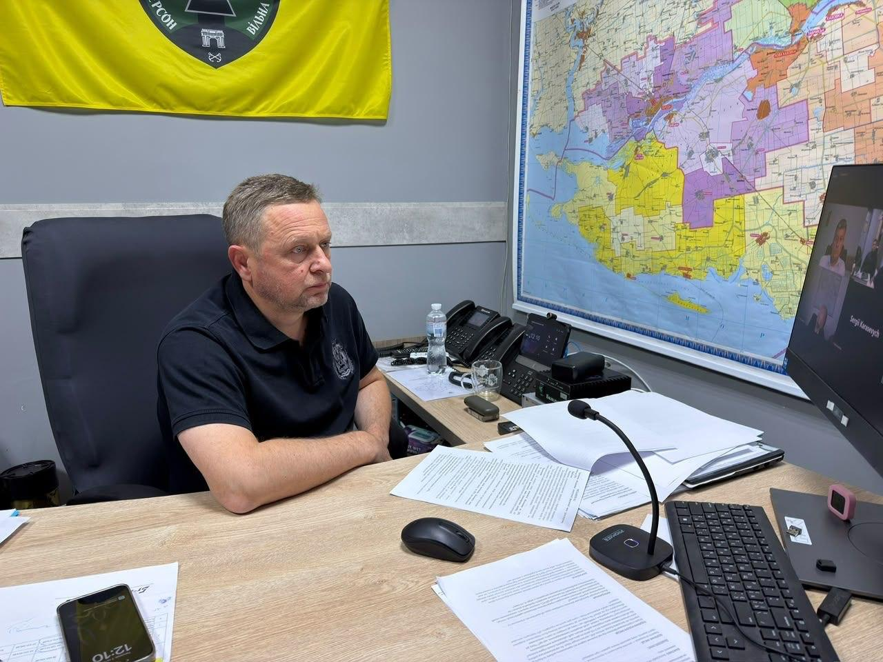 Перший заступник начальника Херсонської ОВА Дмитро Бутрій сидить у робочому кріслі за столом у кабінеті під час онлайн-наради із заступником Міністра аграрної політики та продовольства України Віталієм Головнею щодо спеціальних умов вивезення врожаю з прифронтових територій. Посадовець вбраний у чорну футболку-поло та зосереджено дивиться на великий монітор комп'ютера праворуч, де через відеозв'язок підключено інших учасників зустрічі. На столі перед ним розкладені робочі документи, комп'ютерна периферія, настільний мікрофон, телефони та стаціонарні засоби зв'язку. На стіні позаду Бутрія закріплено жовту частину державного прапора України із написом «ВІЛЬНА», а праворуч розташована велика детальна мапа Херсонської області з кольоровим зонуванням громад.