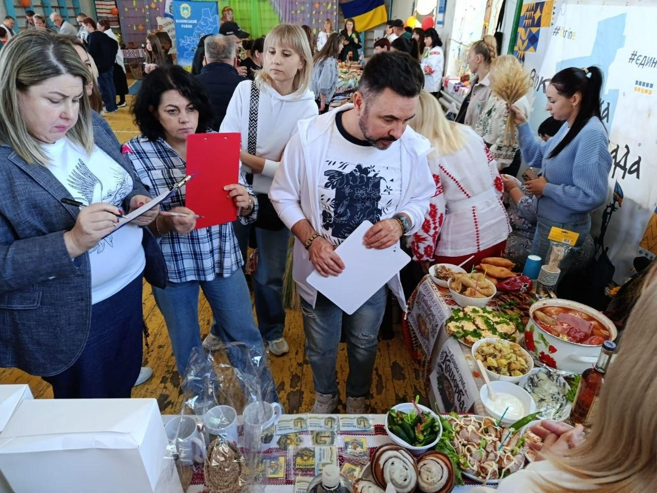 Директорка Департаменту реалізації гуманітарної політики Херсонської ОДА Віолетта Царькова (у картованій сорочці з червоною папкою-планшетом) та заступник директора Департаменту – начальник управління культури Андрій Ярьоменко (у білій сорочці з білим планшетом) працюють у складі професійного журі під час культурно-гастрономічного заходу «Смакуємо у рідному селі» у Калинівській громаді. Разом із колегами вони оцінюють кулінарні презентації громад та роблять нотатки. Поруч на столах представлено розмаїття традиційних українських страв Херсонщини: вареники, голубці, пиріжки, м'ясні нарізки, соління та випічка. На задньому плані видно інших учасників та відвідувачів благодійного ярмарку у святково прикрашеному залі.