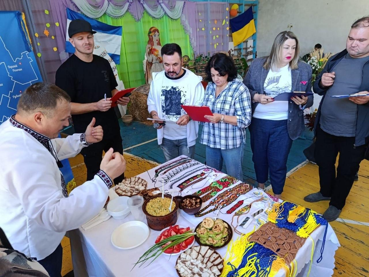 Директорка Департаменту реалізації гуманітарної політики Херсонської ОДА Віолетта Царькова (в центрі у картованій сорочці з червоним планшетом) та заступник директора Департаменту – начальник управління культури Андрій Ярьоменко (ліворуч від неї у білій кофті з капюшоном) разом із колегами по професійному журі оцінюють конкурсний стіл однієї з громад під час заходу «Смакуємо у рідному селі». Ліворуч перед ними стоїть чоловік у вишиванці, який емоційно презентує виставлені страви та вироби, показуючи піднятий догори великий палець. На білій скатертині столу розкладені традиційне сало з проріззю, козацький куліш, свіжі томати, кабачки, а також різноманітні вишиті та плетені сувеніри ручної роботи у синьо-жовтих кольорах. Захід проходить у спортивній залі з тематичними декораціями, святковою фотозоною та українськими прапорами.