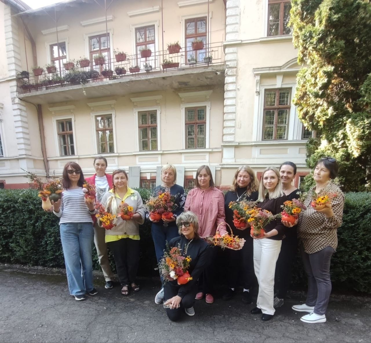 Учасниці заняття з арттерапевтичним компонентом «Осіння гармонія: флористичний гарбуз» від Координаційного центру «Вільні разом» позують для спільного фото на подвір'ї Ботанічного саду Чернівецького національного університету імені Юрія Федьковича. Жінки стоять та сидять у ряду на тлі старовинної світлої будівлі з балконом та вікнами, а також густого зеленого живоплоту й туї. Майже кожна учасниця тримає в руках свій власноруч створений яскравий осінній букет із квітів, сухоцвітів та листя, де замість вази використано декоративний гарбуз, а одна з жінок попереду тримає плетений кошик із квітами. Світлина зафіксувала творчий захід, спрямований на соціальну та психологічну підтримку переселенців з Херсонщини у Чернівцях.
