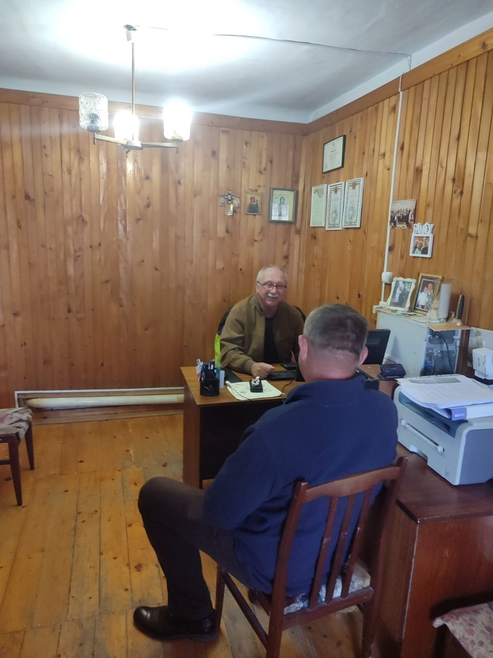 Адміністратор ЦНАПу та юрист-волонтер координаційного центру В.І. Чебан здійснює прийом переселенця з Херсонщини у Чернівцях. Правник літнього віку в окулярах та коричневій куртці сидить за робочим столом, усміхається та спілкується з громадянином, який сидить на стільці спиною до кадру. Робоче місце облаштоване в кабінеті з дерев'яним оздобленням стін та підлоги, де на столі лежать документи, а поруч на тумбі встановлено принтер. На стінах позаду юриста розвішані грамоти, дипломи та сімейні фотографії в рамках. Світлина ілюструє процес надання адміністративних та юридичних послуг для підтримки внутрішньо переміщених осіб від Координаційного центру «Вільні разом».