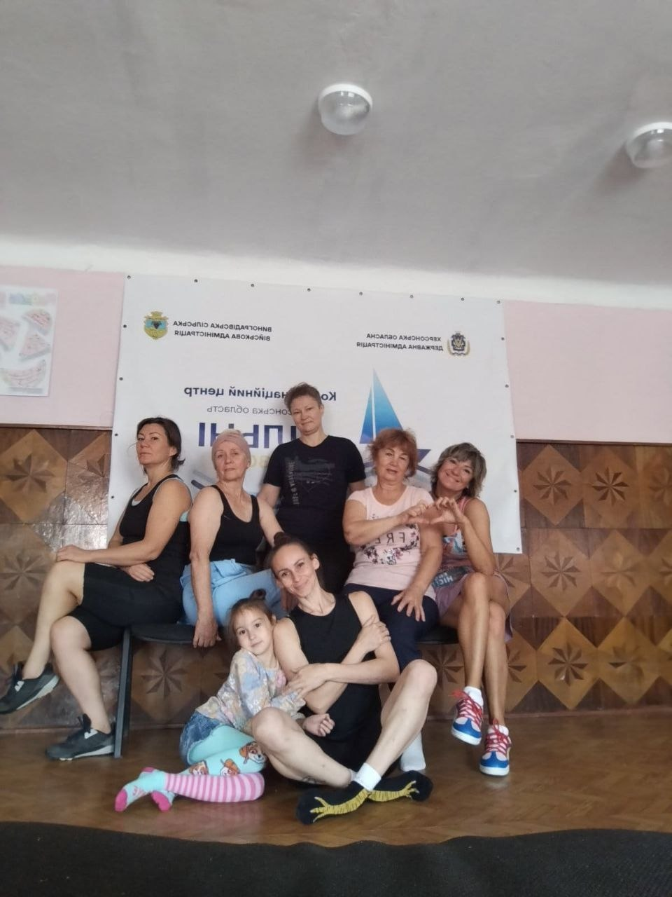 Група жінок-учасниць та маленька дівчинка разом із фітнес-тренеркою Іриною Котовою позують для спільного фото після щотижневого тренування «Будь сильною» від Координаційного центру «Вільні разом» у Чернівцях. Жінки у спортивному одязі посміхаються на тлі банера центру з дзеркально відображеними написами Херсонської ОВА та Виноградівської СВА, при цьому дві учасниці праворуч показують руками жест у формі серця. Тренерка з дитиною розмістилися на підлозі на передньому плані, а світлина відображає момент психоемоційного розвантаження та підтримки здоров'я переселенок з Херсонщини.