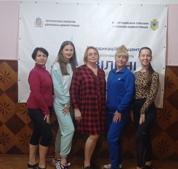 П'ятеро жінок-переселенок з Херсонщини позують для спільного фото на тлі білого банера Координаційного центру «Вільні разом» у Чернівцях. Вони посміхаються після заняття із сальси та бачати, яке пройшло в межах жіночого клубу «Рідна» за сприяння Палацу молоді та студентів Херсонської обласної ради. На банері позаду учасниць видно написи «Херсонська обласна державна адміністрація» та «Виноградівська сільська військова адміністрація». Світлина зафіксувала момент танцювального та психоемоційного розвантаження внутрішньо переміщених осіб.