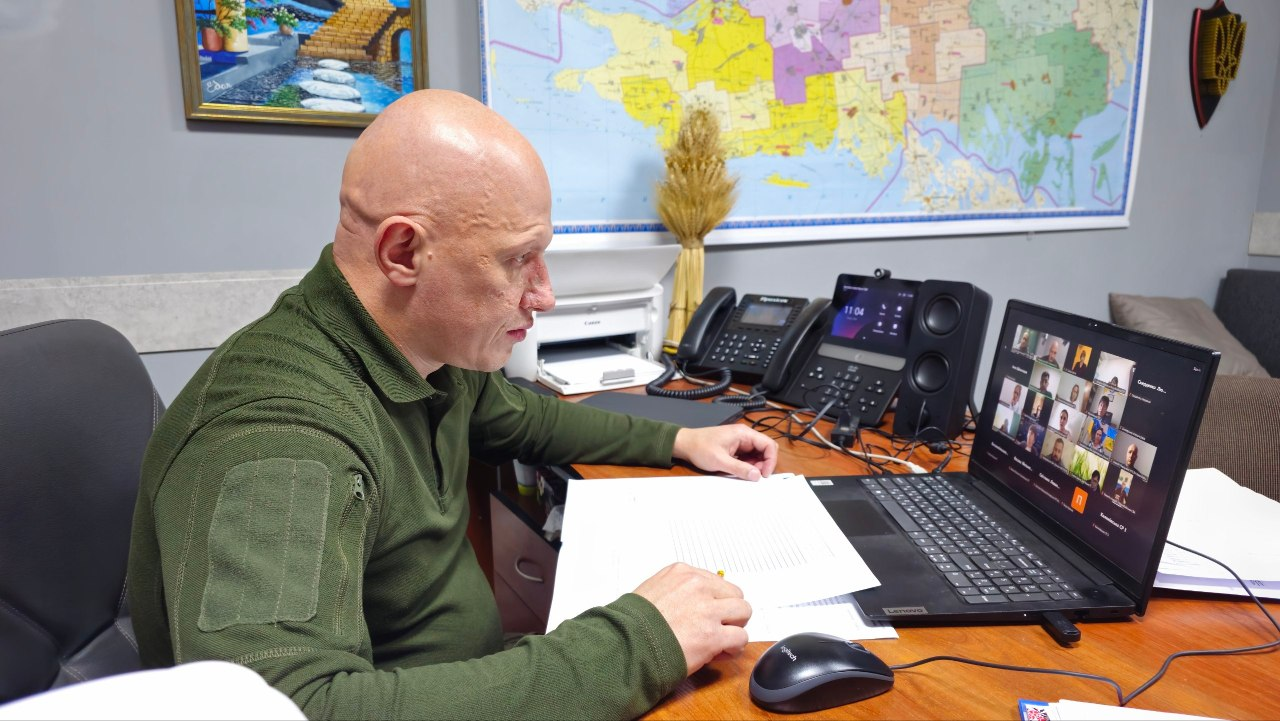 Світлина робочого процесу, на якій заступник начальника Херсонської ОВА Євгеній Ігнатенко проводить онлайн-нараду в кабінеті щодо виплат у межах державної ініціативи «Зимова підтримка» та забезпечення жителів деокупованих громад твердим паливом, де чоловік військової статури із голеною головою, одягнений у флісову кофту хакі з велкро-панеллю на рукаві, сидить за робочим столом у профіль і дивиться на екран ноутбука із запущеною відеоконференцією з багатьма учасниками, тримаючи руку на паперових документах; на столі перед ним також розміщені олівець, комп'ютерна миша, стаціонарний та ІР-телефон, а на сірій стіні позаду висять велика кольорова мапа Херсонської області, декоративний сніп пшениці, картина з пейзажем та герб України на темному щиті.